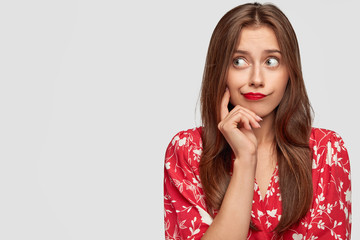 Photo of comely European female wears red lipstick, flower print blouse, keeps finger on cheek and looks thoughtfully aside, poses against white background with copy space for your promotion