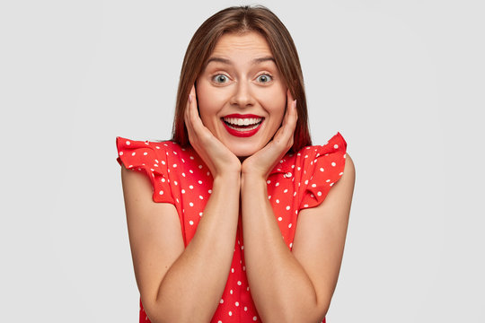 Image of easily moved happy young European woman has broad joyful smile, keeps hands on cheeks, looks happily at camera, dressed in stylish polka dot blouse, stands against white background.