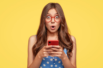 Surprised young female model holds modern cell phone, reads sudden news on internet web page, surfes in social networks, poses against yellow background. People, emotions, technology, lifestyle - obrazy, fototapety, plakaty