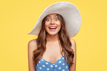 Indoor shot of beautiful joyful traveller has overjoyed expression, long wavy hair, wears summer hat and casual dress, notices real paradise in front, stands against yellow wall. People and fashion