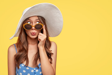 Stylish woman with surprised mysterious expression, looks asdie, wears stylish summer shades, hat and polka dot blouse, isolated over yellow background with copy space for your text. Relaxation - obrazy, fototapety, plakaty