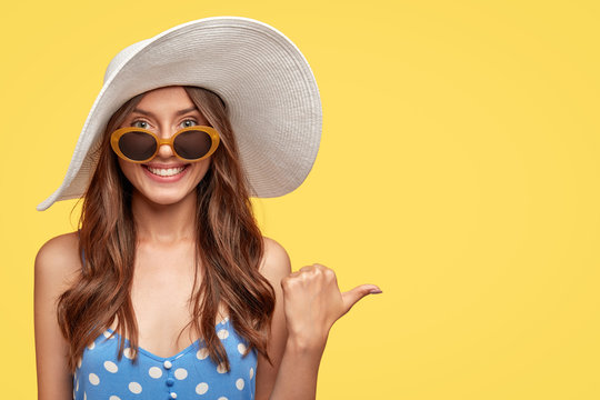 Summer time concept. Cheerful beautiful brunette woman in stylish sunglasses and hat, has friendly smile, points with thumb aside, shows blank space for your advertisement or promotional text