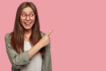 Indoor shot of attractive Caucasian female with happy expression, warm smile, points with index finger at upper right corner, isolated over pink background, dressed casually. Look there, its awesome!