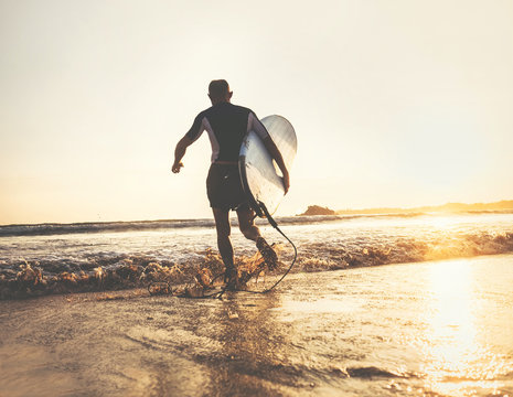 Surfer with surfboard runs in ocean waves, sunset time. Active lifestyle concept