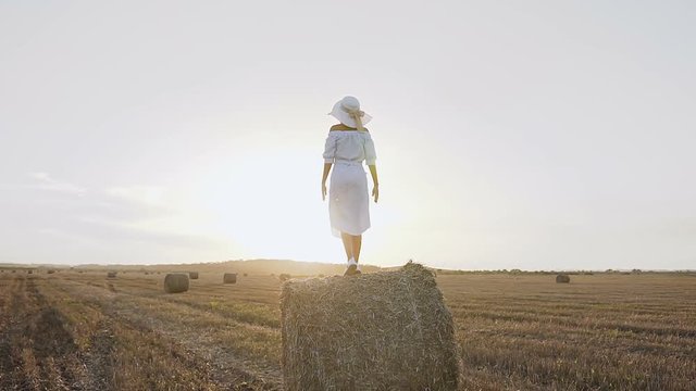 Back view. Young girl in dress and big hat standing on haystack with outspread hands have fun at sunset in the field
