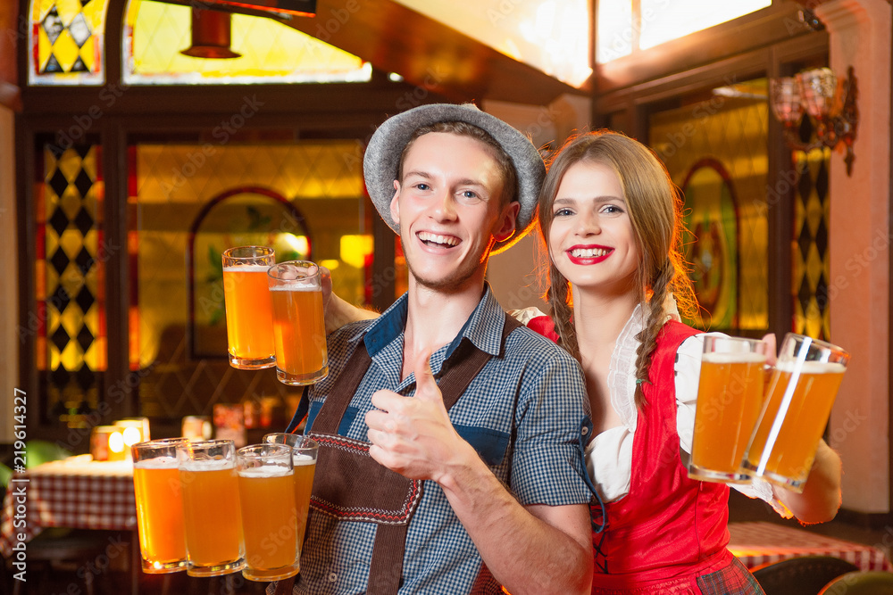 Wall mural Cheerful man and girl waiters in national costumes at the celebration octoberfest hold in their hands a lot of beer mugs.