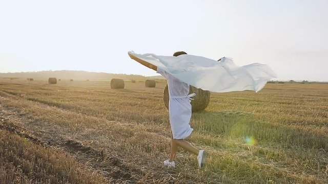 Back view. Beautiful girl in long dress having fun outdoors in the field. Happy brunette woman walking with scarf in the field with straw bales and enjoying nature