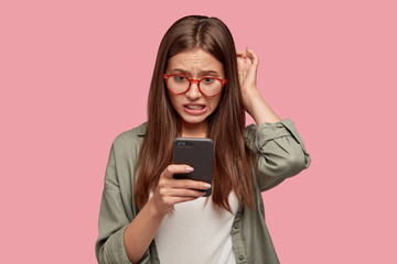 Nervous puzzled woman scratches head in bewilderment, reads bad news at internet, uses modern cell...
