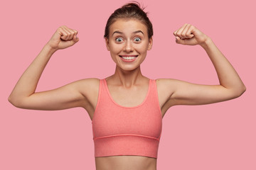 Sporty energetic athletic woman wears pink sports top smiles as shows her biceps, likes sport and...