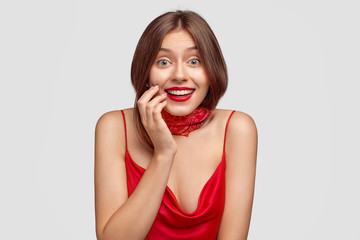 Lively model with positive expression, wears casual red dress and bandana on neck, looks exultant, rejoices unforgettable moments in life, uses cosmetics, poses alone against white background