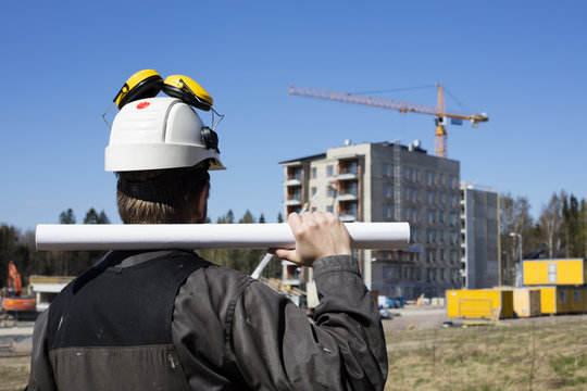 Construction worker in dirty overalls and a white helmet in Finland. In his hand he has drawings. In the background is out of focus in the construction site. 