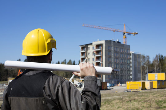 Construction Worker In Dirty Overalls And A Yellow Helmet In Finland. In His Hand He Has Drawings. In The Background Is Out Of Focus In The Construction Site. 