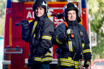 Photo of two young firemen with axes in hands near fire engine