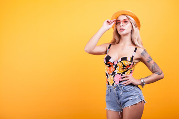 Young sexy slim tanned woman in swimsuit with sunglasses and hat over yellow background