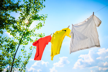 Image of three different colored T-shirts hanging on ropes against blue sky with tree tops.