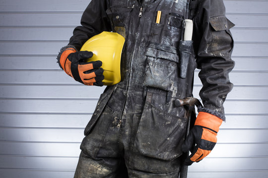 Construction worker in dirty overalls in Finland. The laborer have orange gloves, yellow helmet and hammer. Background out of focus and illuminated with flash. 
