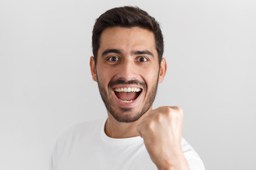 Daylight portrait of young passionate man isolated on gray background wearing casual t-shirt smiling and laughing in excitement while supporting team or celebrating victory in competition