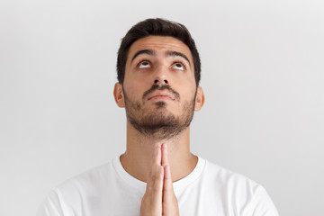 Obraz premium Indoor close up daylight picture of young man isolated on gray background, dressed in white t-shirt, praying with hands pressed together, looking upwards as if asking god for blessing