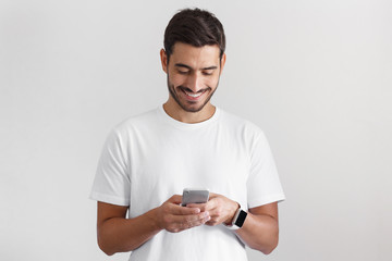 Daylight portrait of young european caucasian man isolated on gray background wearing white t-shirt standing in front of camera, looking attentively with smile at screen of smartphone he is holding