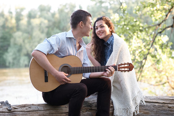 young couple sitting on a log by the river and playing guitar, summer nature, bright sunlight, shadows and green leaves, romantic feelings