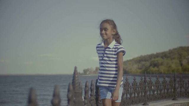 Portrait of positive cute elementary age african american girl in casual clothes walking along parapet holding fence while enjoying view during summer vacation trip at seaside.
