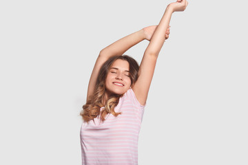 Happy relaxed Caucasian woman stretches after awakening, being in good mood as saw pleasant dreams, dressed in nightwear, ready to start new day, stands against white background. Rest concept