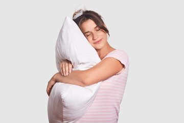 Photo of relaxed young female has sweet dreams, embraces soft pillow, wears nightclothes and has...