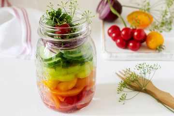 Salad of vegetables with herbs in a glass jar and fresh vegetables on a table.