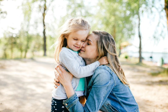 Happy smiling mother hugging her lovely little daughter outdoor. Lifestyle family. Adult cheerful female parent playing with her beautiful emotional child at nature in summer. Positive people faces.