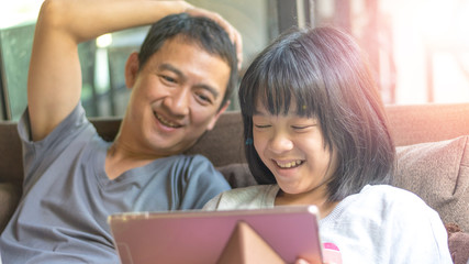 Father and daughter activity technology together concept. Asia kid girl reading e-book or playing game on computer tablet while dad watching as parental guidance for PG13.