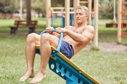 Outdoor shot of attractive fit healthy young male works abs on special sport equipment, wears shorts, shows his mans power, has cardio training in park in morning. People, recreation concept