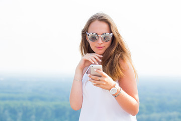 A beautiful young woman in sunglasses looks at the smartphone with a smile, reads the message, goes to the Internet