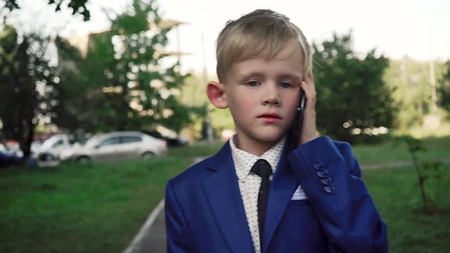 Small boy with a serious face in a business suit is walking down the street and talking on the phone