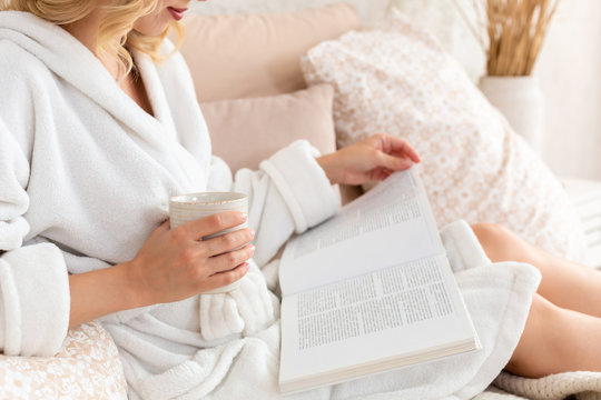 Young Woman In White Terry Robe Is Drinking Coffee And Reading Magazine Or Book In Bedroom.