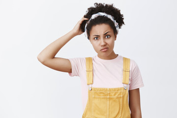 I am confused. Portrait of unsure doubtful cute African American young woman in yellow overalls and headband, scracthing head, pursing lips and frowning while thinking, being questioned and uncertain