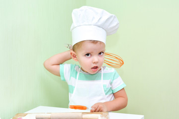 A little cute boy holds an orange whisk for eggs and he wants to take off the white cap of the chef