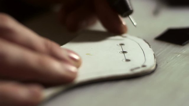 Handmade tailoring in shoes production. Close up leather craftsman hands working with leather sketch. Artisan man making holes in leather blank with awl. Design process leather goods in industry