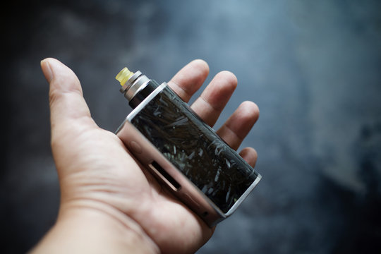 high end black carbon fiber in clear resin regulated box mods with rebuildable dripping atomizer and ultem drip tip in hand on dark grey texture background, vaporizer equipment, selective focus