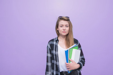 Portrait of a student girl standing on a purple background with books and notebooks in her hands and looking into the camera. Serious student is isolated on a purple background.