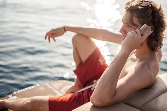 Caucasian Man Traveler Floating On Motorboat Along Sea Coast With Hotels And Urban Scape And Chatting With A Friend On Smartphone. Summer Vacation At Seaside.