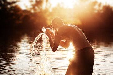 a young man is standing in a river and washing his face against the sunset.