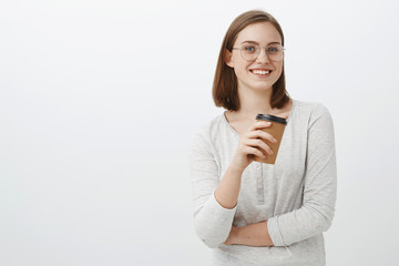 Creative happy and enthusiastic charming female office worker having break standing in cafe over gray background holding paper cup of coffee talking amused with coworker smiling at camera