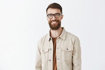 Waist-up shot of friendly-looking pleased handsome caucasian bearded guy with moustache smiling satisfied and happy standing joyfully over gray background in beige jacket