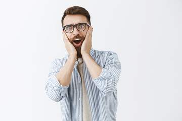 Waist-up shot of impressed and surprised sociable good-looking male with long beard and moustache holding palms on cheeks and staring at camera hearing shocking rumor over gray background