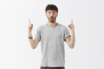 Naklejka premium Guy easy to impress. Portrait of emotive enthusiastic caucasian dark-haired young man with beard and stylish dark hairstyle folding lips with joy, raising index fingers and pointing up with amazement
