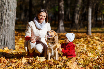 Fototapeta premium Cute, happy, white boy in red shirt smiling and playing with dog among yellow leaves. Little child having fun with his mom in autumn park. Concept of friendship between kids and pets, happy family