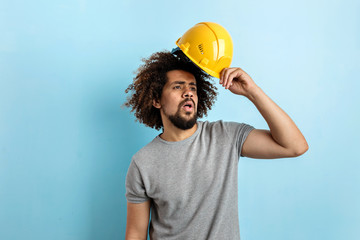 A curly-headed handsome man wearing a gray T-shirt is standing with a safety helmet and looking sideways over the blue background.
