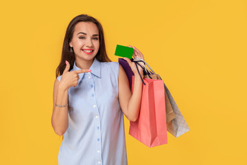 Fototapeta na wymiar Trendy woman 20s in dress with long brown hair smiling while holding different shopping packages and card in hands isolated over yellow background