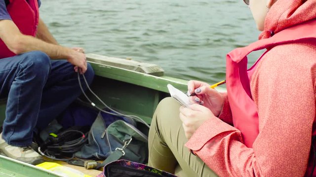 Close up man and woman environmental scientists in a research boat. Man measuring pH of the lake water, woman writing result in a diary