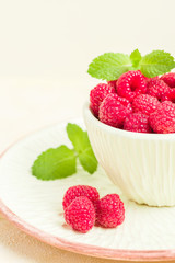 Ripe raspberries with green mint leaves in cup and saucer on pastel yellow background.